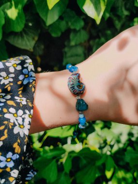 Mavi taşlar, kadının bileğindeki kyanit apatit boncuklu bilezik. Yeşil yapraklar arka plan, güneş ışığı, gölgeler, yaz. Yarı değerli taş makrome girdap bilezik, mücevher, aksesuar, bijouterie