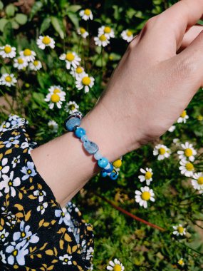 Mavi taşlar kyanit aquamarine apatite. Kadının bileğindeki boncuklu bilezik. Papatya çiçeklerinin arasına yakın. Güneşli bir yaz günü. Yarı değerli taş makrome shamballa bilezik, mücevher, aksesuar, bijouterie