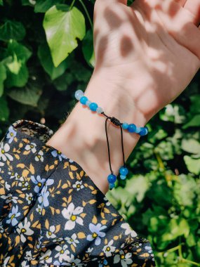 Mavi taşlar, kadının bileğindeki kyanit apatit boncuklu bilezik. Yeşil yapraklar arka plan, güneş ışığı, gölgeler, yaz. Yarı değerli taş makrome ayarlanabilir kilit bilezik, mücevher, aksesuar. 