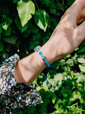 Mavi taşlar, kadının bileğindeki kyanit apatit boncuklu bilezik. Yeşil yapraklar arka plan, güneş ışığı, gölgeler, yaz. Yarı değerli taş makrome girdap bilezik, mücevher, aksesuar, bijouterie