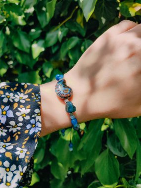 Mavi taşlar, kadının bileğindeki kyanit apatit boncuklu bilezik. Yeşil yapraklar arka plan, güneş ışığı, gölgeler, yaz. Yarı değerli taş makrome girdap bilezik, mücevher, aksesuar, bijouterie