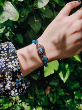 Mavi taşlar, kadının bileğindeki kyanit apatit boncuklu bilezik. Yeşil yapraklar arka plan, güneş ışığı, gölgeler, yaz. Yarı değerli taş makrome girdap bilezik, mücevher, aksesuar, bijouterie