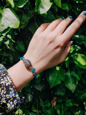 Mavi taşlar, kadının bileğindeki kyanit apatit boncuklu bilezik. Yeşil yapraklar arka plan, güneş ışığı, gölgeler, yaz. Yarı değerli taş makrome girdap bilezik, mücevher, aksesuar, bijouterie