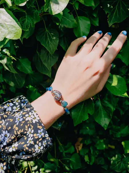 Mavi taşlar, kadının bileğindeki kyanit apatit boncuklu bilezik. Yeşil yapraklar arka plan, güneş ışığı, gölgeler, yaz. Yarı değerli taş makrome girdap bilezik, mücevher, aksesuar, bijouterie