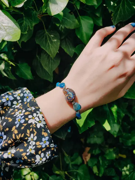 Mavi taşlar, kadının bileğindeki kyanit apatit boncuklu bilezik. Yeşil yapraklar arka plan, güneş ışığı, gölgeler, yaz. Yarı değerli taş makrome girdap bilezik, mücevher, aksesuar, bijouterie