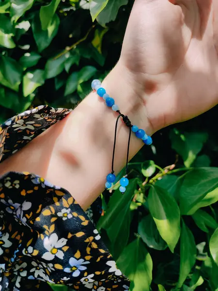 Mavi taşlar, kadının bileğindeki kyanit apatit boncuklu bilezik. Yeşil yapraklar arka plan, güneş ışığı, gölgeler, yaz. Yarı değerli taş makrome ayarlanabilir kilit bilezik, mücevher, aksesuar. 