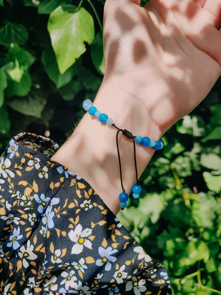 Mavi taşlar, kadının bileğindeki kyanit apatit boncuklu bilezik. Yeşil yapraklar arka plan, güneş ışığı, gölgeler, yaz. Yarı değerli taş makrome ayarlanabilir kilit bilezik, mücevher, aksesuar. 