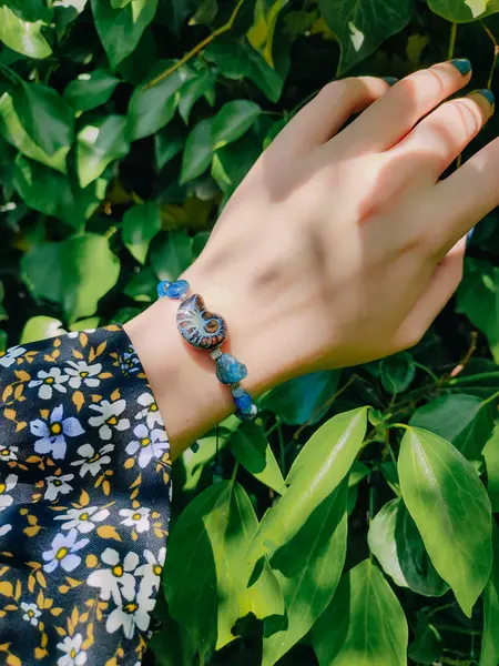 Mavi taşlar, kadının bileğindeki kyanit apatit boncuklu bilezik. Yeşil yapraklar arka plan, güneş ışığı, gölgeler, yaz. Yarı değerli taş makrome girdap bilezik, mücevher, aksesuar, bijouterie