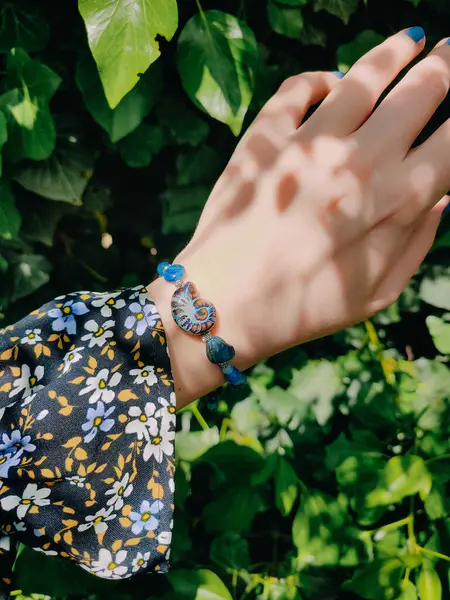 Mavi taşlar, kadının bileğindeki kyanit apatit boncuklu bilezik. Yeşil yapraklar arka plan, güneş ışığı, gölgeler, yaz. Yarı değerli taş makrome girdap bilezik, mücevher, aksesuar, bijouterie