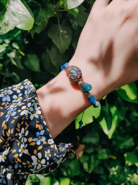 Mavi taşlar, kadının bileğindeki kyanit apatit boncuklu bilezik. Yeşil yapraklar arka plan, güneş ışığı, gölgeler, yaz. Yarı değerli taş makrome girdap bilezik, mücevher, aksesuar, bijouterie