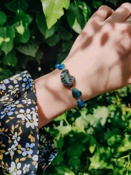 Mavi taşlar, kadının bileğindeki kyanit apatit boncuklu bilezik. Yeşil yapraklar arka plan, güneş ışığı, gölgeler, yaz. Yarı değerli taş makrome girdap bilezik, mücevher, aksesuar, bijouterie