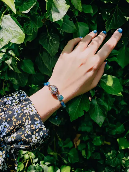 Mavi taşlar, kadının bileğindeki kyanit apatit boncuklu bilezik. Yeşil yapraklar arka plan, güneş ışığı, gölgeler, yaz. Yarı değerli taş makrome girdap bilezik, mücevher, aksesuar, bijouterie