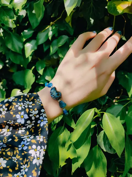 Mavi taşlar, kadının bileğindeki kyanit apatit boncuklu bilezik. Yeşil yapraklar arka plan, güneş ışığı, gölgeler, yaz. Yarı değerli taş makrome girdap bilezik, mücevher, aksesuar, bijouterie