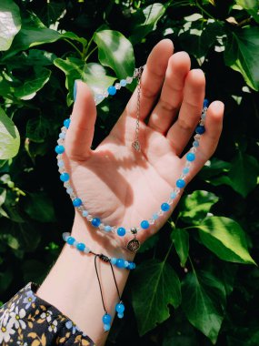 Mavi taşlı, boncuklu apati boncuklu, elinde cazibesi olan bir kadın. Yeşil yapraklar, güneş ışığı ve gölgeler. Doğal değerli taş mücevherler, aksesuar, kolye, bijouterie. 