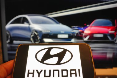 Roosendaal, Hollanda - 6 Nisan 2025: Hyundai logosunu gösteren bir cep telefonu, arka planda Ioniq 6 'nın güncellenmiş ve yüz gerdirilmiş bir resmi.