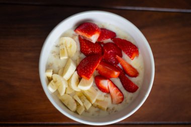 Üstünde taze muz dilimleri ve sulu çilekler olan kremalı yulaf ezmesi olan sağlıklı bir kahvaltı kasesinin üst görüntüsü. Canlı ve besleyici bir yemek fikri sağlıklı beslenmeyi, sabah rutinlerini veya vejetaryen diyet kavramlarını desteklemek için mükemmeldir.