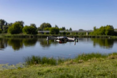 Modern bir dron güneşli bir günde sakin bir nehrin üzerinde süzülür. Yeşil ağaçlar ve açık mavi bir gökyüzü barışçıl bir doğal zemin oluşturur. Hava fotoğrafçılığı kavramı, doğadaki teknoloji ve açık hava yeniliği..