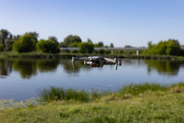 Modern bir dron güneşli bir günde sakin bir nehrin üzerinde süzülür. Yeşil ağaçlar ve açık mavi bir gökyüzü barışçıl bir doğal zemin oluşturur. Hava fotoğrafçılığı kavramı, doğadaki teknoloji ve açık hava yeniliği..