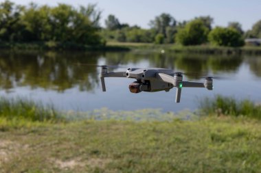 Modern bir dron güneşli bir günde sakin bir nehrin üzerinde süzülür. Yeşil ağaçlar ve açık mavi bir gökyüzü barışçıl bir doğal zemin oluşturur. Hava fotoğrafçılığı kavramı, doğadaki teknoloji ve açık hava yeniliği..