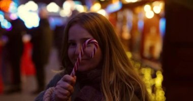 Neşeli bir videoda bir kız Noel pazarında duruyor, gülümsüyor ve neşeyle Noel şekeri sallıyor..