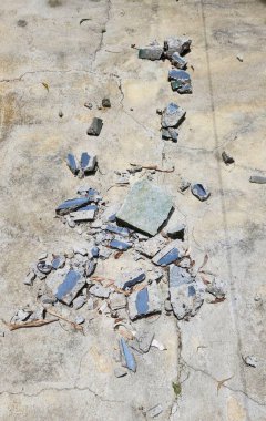 Kırık ve kırık beton kaldırımın detaylı görüntüsü moloz ve molozlar etrafa saçılmış, zaman içinde aşınma ve aşınma gösteriyor, küçük mavi parçalar kentsel bozulmayı sembolize ediyor..