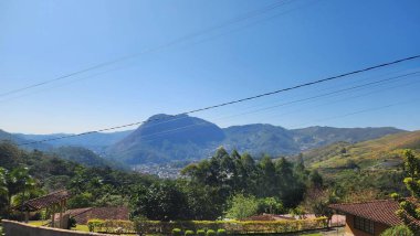 Berrak mavi gökyüzünün altında, yeşil bitki örtüsü, dağınık evler ve elektrik hatlarıyla ön planda yavaşça çaprazlamasına uzanan resim gibi bir dağ sırasını yakalayan sakin bir manzara..