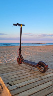 Günbatımında kumlu sahile ve okyanusa bakan ahşap bir kaldırıma park etmiş modern bir elektrikli scooter. Arka planda renkli bir gökyüzü var..