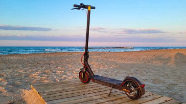 Günbatımında kumlu sahilde, ahşap bir kaldırımda modern bir elektrikli scooter, sakin okyanus dalgaları ve arka planda renkli bir gökyüzü..