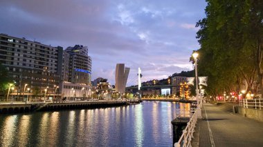İspanya 'nın Bilbao şehrinin güzel bir akşam manzarası. Şehir ışıkları nehri yansıtıyor. Modern mimari ve dramatik bir gökyüzü var..
