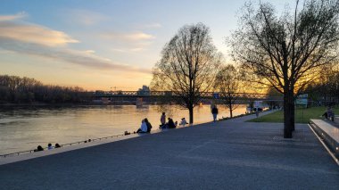 Bratislava, Slovakya 'daki Tuna Nehri' nin üzerinde güzel bir günbatımı. Nehir kenarındaki gezinti alanında oturan insanlar, altın akşam atmosferinin tadını çıkarıyorlar..