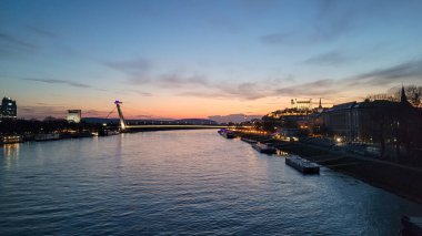 Bratislava, Slovakya 'daki Tuna Nehri üzerinde büyüleyici bir alacakaranlık manzarası, aydınlatılmış UFO Köprüsü, Bratislava Kalesi ve suyun üzerine yansıyan şehir ışıkları.