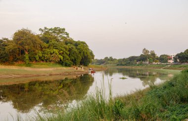 Sahil boyunca yemyeşil ağaçlarla dolu huzurlu bir nehir manzarası ve gökyüzünü yansıtan sakin bir su yüzeyi sakin bir atmosfer yaratıyor..