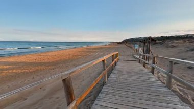 Sakin, ahşap bir patika gün batımında kumlu bir sahile çıkar. Sakin okyanus dalgaları yavaşça kıyıya vurur.