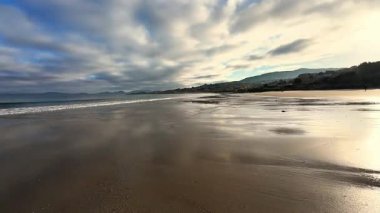 Huzurlu ve sakin bir sahil sahnesi canlı yansımaları ve karamsar bulutları gökyüzünde sergiliyor.