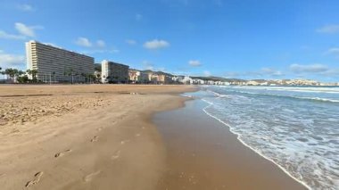 Sakin bir sahil sahnesi. Açık bir gökyüzü altında altın kum, yumuşak dalgalar ve kıyı mimarisi sergileniyor..