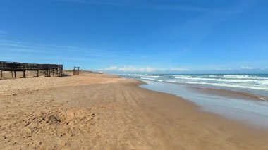 Huzurlu ve pitoresk bir plaj sahnesinde yumuşak altın kum, yumuşak dalgalar ve açık mavi bir gökyüzü sergilenir.