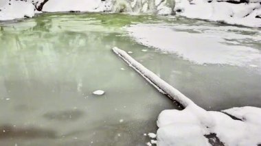 Huzurlu ve pitoresk bir kış manzarası kısmen donmuş bir su kütlesini karla kaplı bir kıyıda sergiliyor.