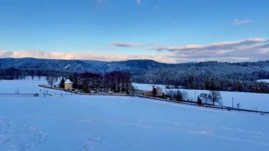 Parlak mavi gökyüzünün altında karla kaplı tarlalar, ağaçlar ve yuvarlanan tepelerin yer aldığı sakin bir kış manzarası..