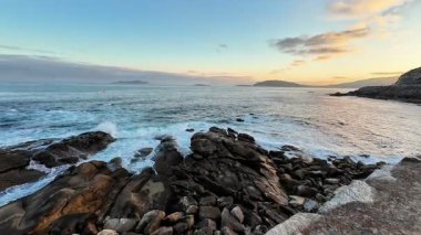 Gün batımında güzel, sakin bir kıyı manzarası, dalgaların ritmik bir şekilde kayalık bir kıyıya çarpması.