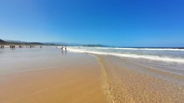 Sakin bir sahil sahnesi berrak mavi gökyüzünü ve kumlu sahilde hafifçe süzülen yumuşak okyanus dalgalarını gösteriyor.