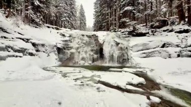 Kar dolu ağaçlar tarafından zarif çerçevelenmiş nefes kesici bir kış şelalesi inanılmaz sakin bir manzara yaratıyor.