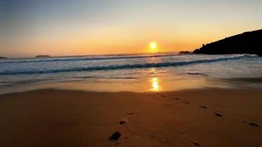 Gün batımında sakin ve huzurlu bir sahil sahnesi. Suya doğru giden yumuşak dalgalar ve ayak izleri sergiliyor.