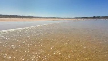 Sakin bir sahil sahnesi, berrak mavi gökyüzü altında yumuşak dalgalar, rahatlamak ve gevşemek için mükemmel.