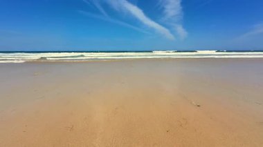 Huzurlu ve sakin bir sahil sahnesi yumuşak altın kumlar ve parlak mavi gökyüzünün altında yumuşak okyanus dalgaları.