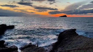 Geniş bir okyanus üzerinde nefes kesici ve renkli bir gün batımı manzarası, güzel bir şekilde sakin dalgaları ve kayalık kıyıları yakalıyor.