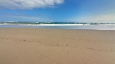Görkemli bir plaj sahnesi. Altın kum, yumuşak dalgalar ve yukarıda parlak mavi bir gökyüzü.