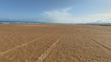 Yumuşak altın kum, yumuşak dingin dalgalar ve yukarıda canlı mavi bir gökyüzü içeren sakin bir sahil sahnesi.