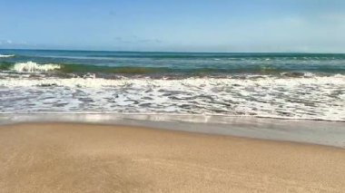 Huzurlu ve sakin bir sahil sahnesi berrak mavi gökyüzünün altında yumuşak, sıcak kum ve nazik okyanus dalgalarını sergiliyor.