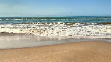 Huzurlu ve sakin bir sahne, okyanus dalgalarının parlak bir gökyüzünün altındaki kumlu güzel bir sahile nazikçe çarptığını tasvir eder.