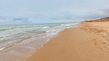 Güzel, sakin bir sahil sahnesi yumuşak altın kum ve yumuşak okyanus dalgalarını sergiliyor. Hepsi bulutlu bir gökyüzünün altında.